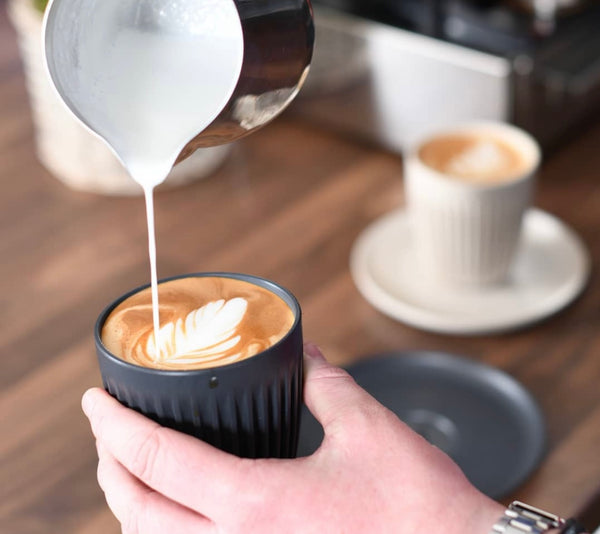 barista pouring flat white coffee into black sustainable huskee coffee cup